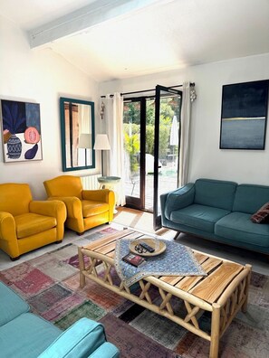 Un salon avec une table basse en bois, deux fauteuils jaunes, un canapé turquoise et une grande fenêtre donnant sur un paysage verdoyant.
