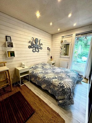 Un dormitorio con cama, escritorio, silla, espejo y ventana con vistas a la vegetación.
