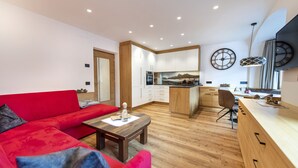 A modern living room with a red sofa, wooden coffee table, and a kitchen area with wooden cabinets and a built-in island. - Apartments Fina 158A (Ortisei)
