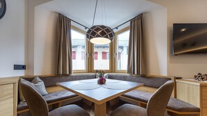 A dining area with a wooden table and bench seating, large windows with curtains, and a flat-screen TV on the wall. - Apartments Fina 158A (Ortisei)