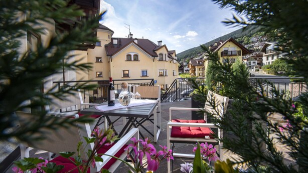 A terrace with outdoor seating, a table with a bottle and glasses, and a view of a hillside village. - Apartments Fina 158A (Ortisei)