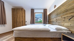 A bedroom with a wooden headboard, a large window with curtains, and a view of buildings outside. - Apartments Fina 158A (Ortisei)