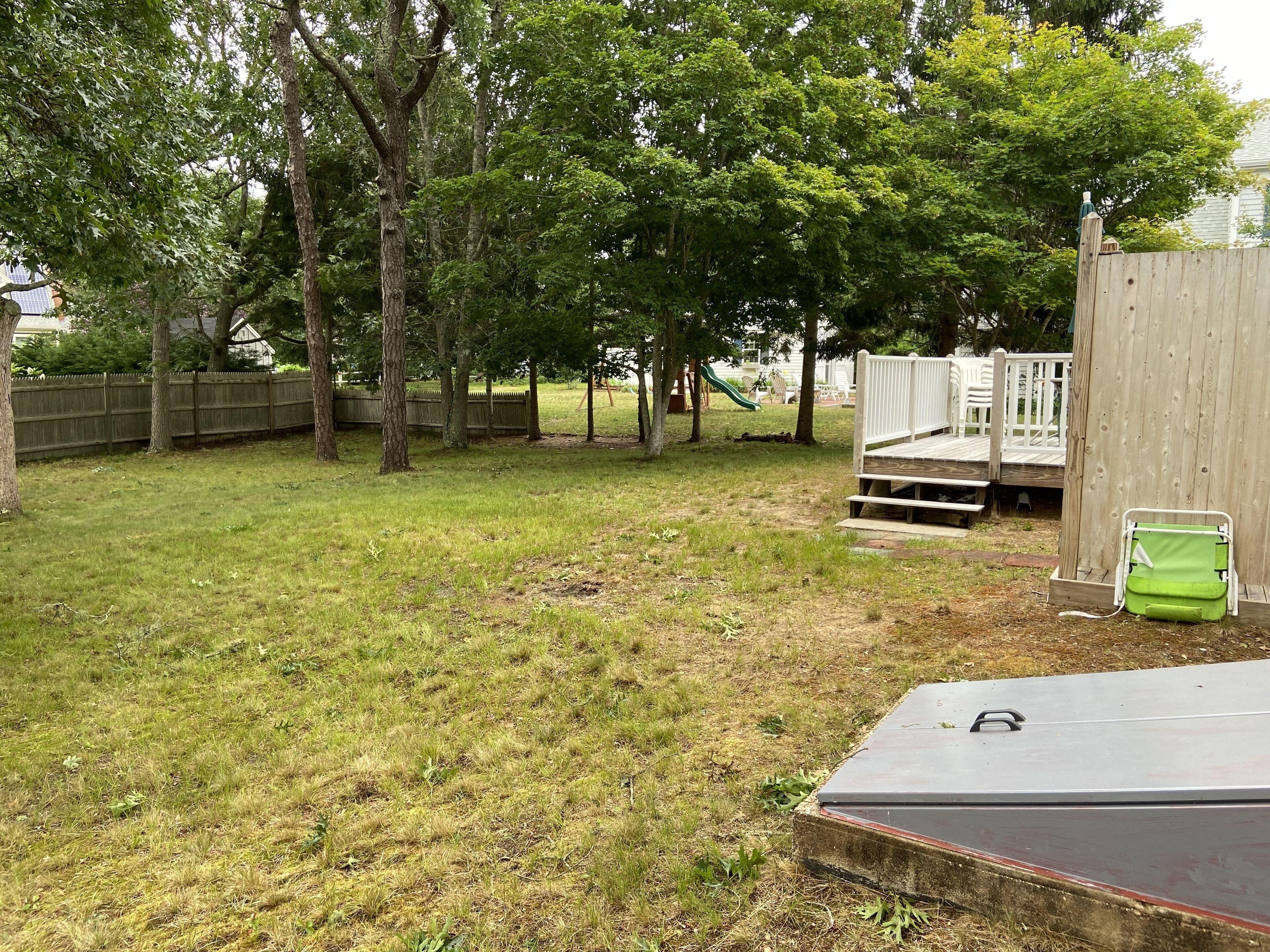 A backyard with a wooden deck, a green slide, and a fenced area with trees.