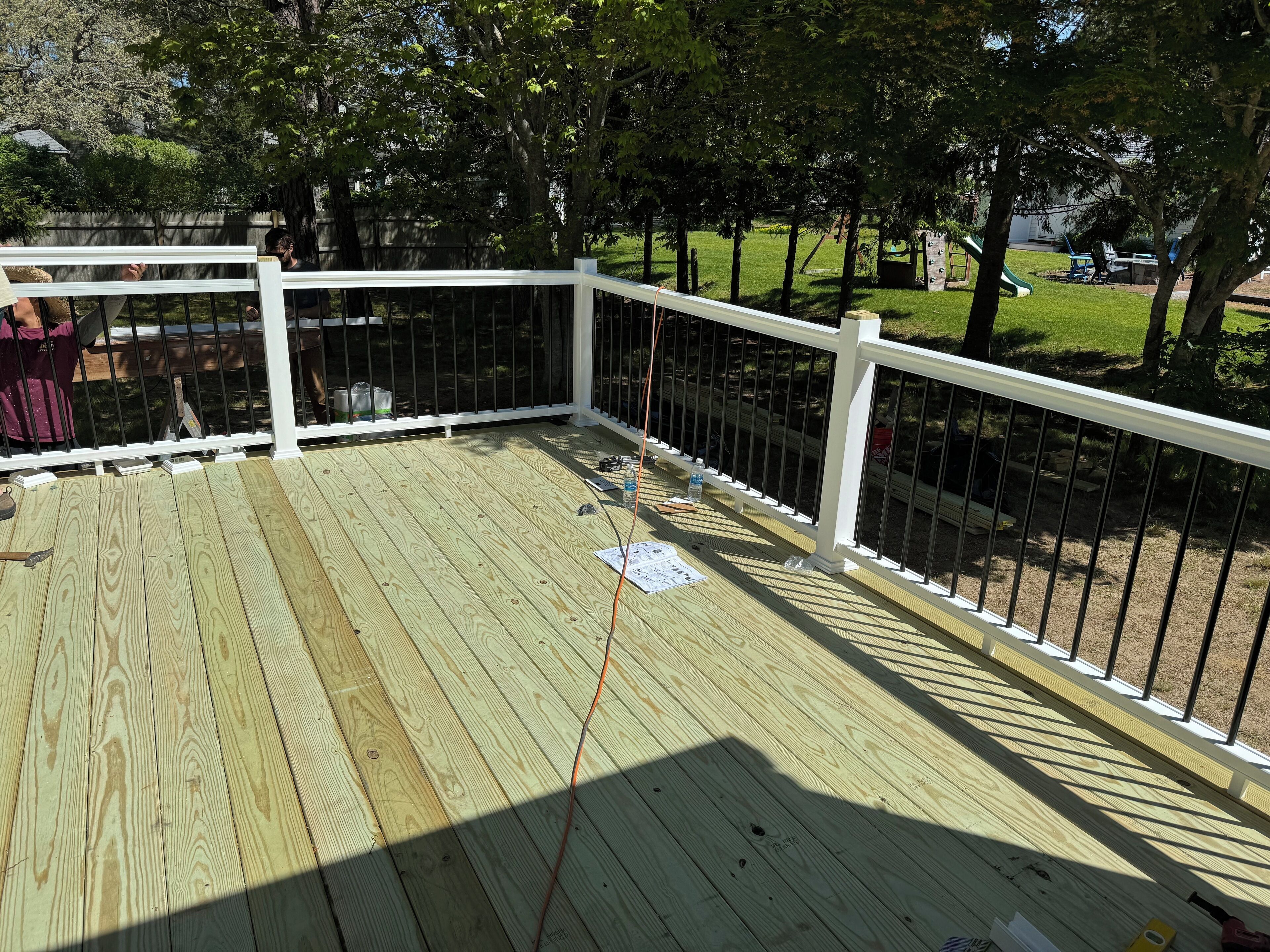 Un patio in legno con ringhiere bianche, circondato da alberi e un'area erbosa con attrezzature da gioco.