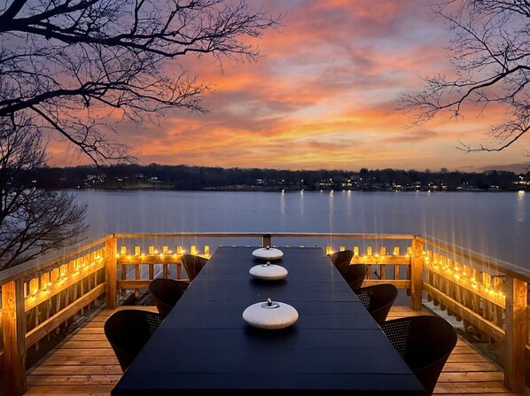 A deck with a long table, chairs, and string lights, overlooking a body of water with a sunset sky.
