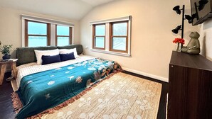 A bedroom with a bed, two windows, a nightstand, a lamp, and a wall-mounted shelf with decorative items.