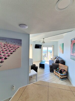 A living room with a large beach-themed painting, a ceiling fan, and a wooden crate with colorful items. - WOW!!!!, ON BEACH, 7TH FL, AMAZING BAY and FIREWORKS VIEW! SPI License 20232542 (South Padre Island)