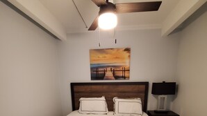 Une chambre avec un lit en bois, un ventilateur de plafond, une table de chevet avec une lampe et un tableau représentant un coucher de soleil sur une jetée.