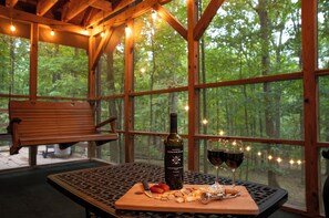 A wooden bench and a table with a bottle of wine and glasses, set against a forest backdrop through large windows. - Woods, River, Relaxation- Berkley Springs (Great Cacapon)