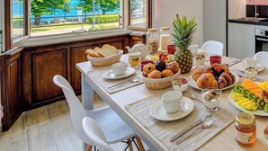 Una mesa de desayuno con diversos alimentos, vistas a árboles y una masa de agua a través de amplias ventanas, y una cocina moderna al fondo.