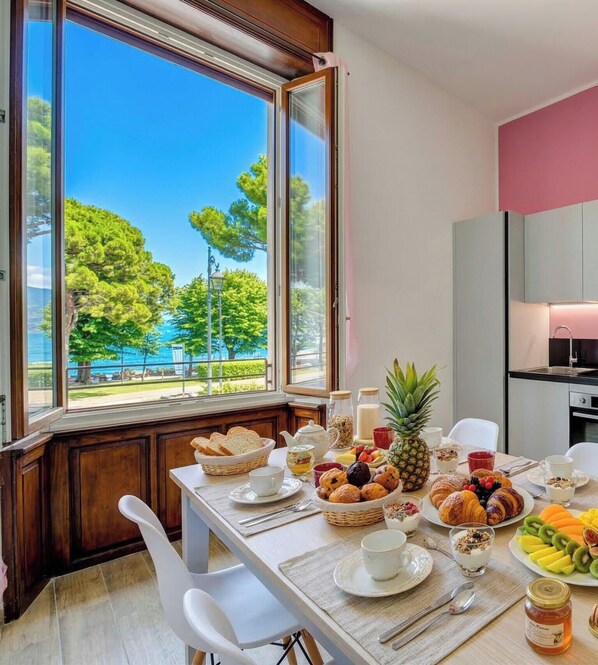 A breakfast table with a variety of foods, a view of trees and a body of water through large windows, and a modern kitchen in the background. - Desenzanoloft Palazzo Visconti - Garda Chiaretto (Desenzano del Garda)