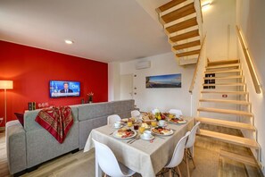 A dining area with a table set for breakfast, a sofa, a TV displaying a news channel, and a staircase.