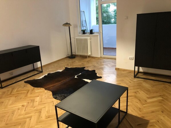Um quarto com piso de madeira em padrão herringbone, uma mesa preta, um armário preto e um sofá preto. Há um tapete de couro preto no chão.