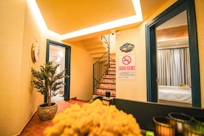 A hotel hallway with a staircase, a potted plant, a bed with a curtain, and a no-smoking sign. - PetraAda Bozcaada (Bozcaada)
