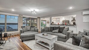 Amplia sala de estar con un ventanal que da a una masa de agua, una chimenea y una zona de cocina con armarios blancos.