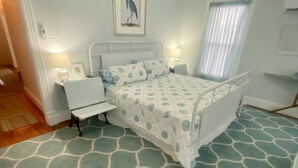 Un dormitorio con cama, mesitas de noche, una mesita auxiliar, un cuadro enmarcado en la pared y una ventana con cortinas.