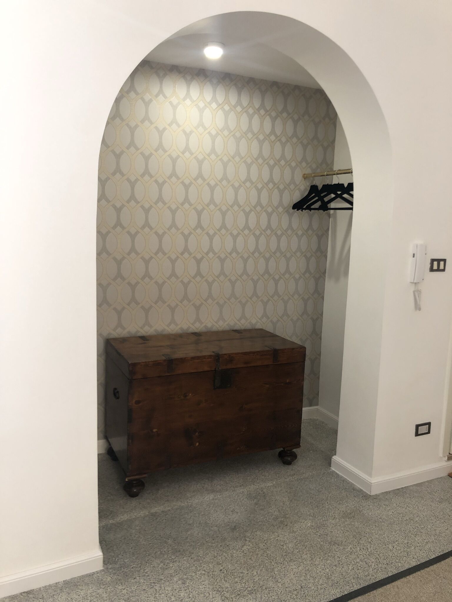 A small room with a patterned wallpaper, a wooden chest of drawers, and a coat rack.
