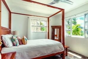 Una cama con dosel, con una colcha blanca, cabecera de madera y varios almohadones. Se ve una ventana con vistas a zonas verdes.