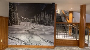 Um corredor com um grande painel decorativo na parede retratando um caminho nevado nas montanhas, lambris de madeira e uma escadaria com corrimãos de metal.