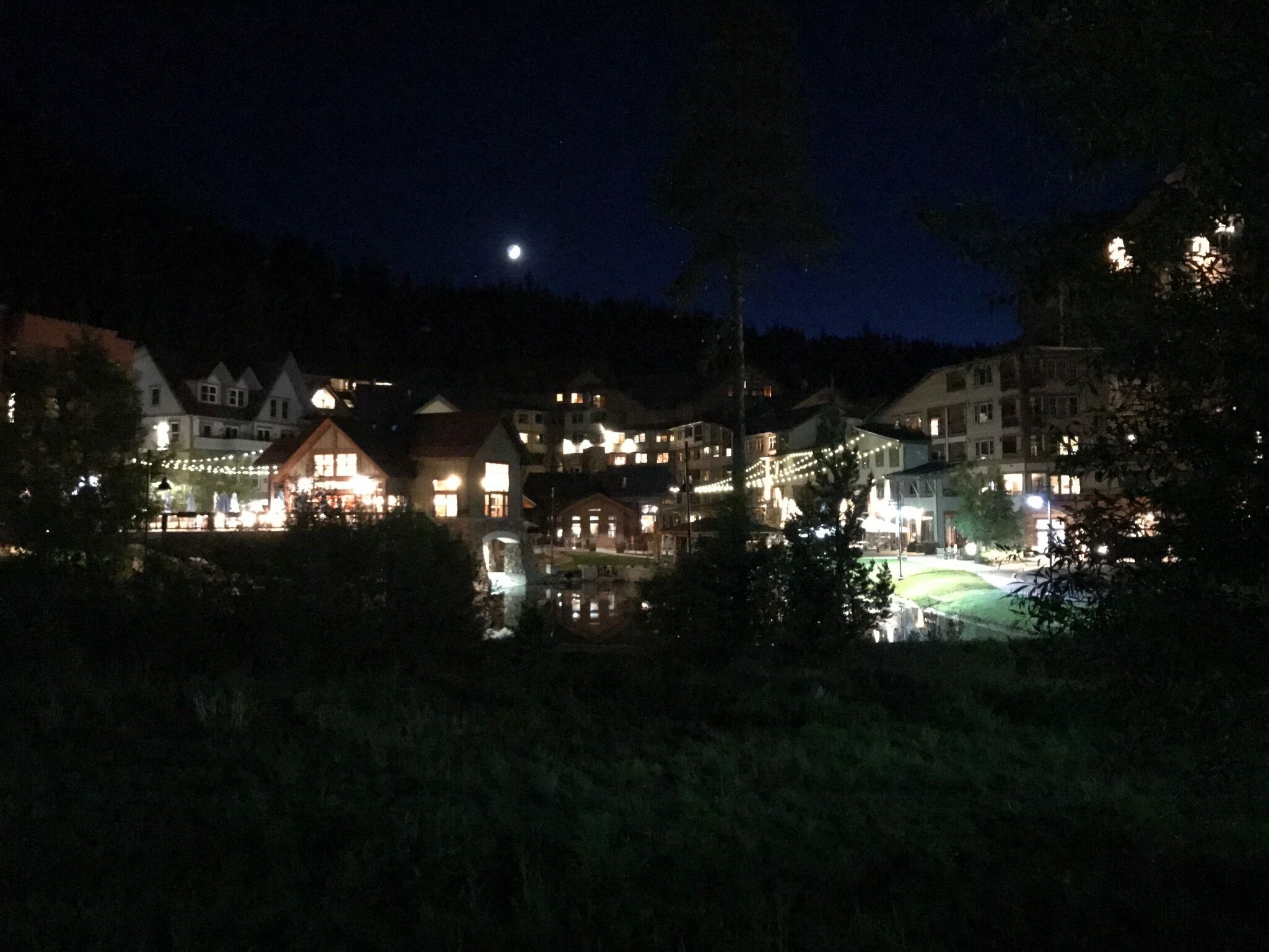 A nighttime view of a lakeside village with illuminated buildings, a visible moon, and a reflection on the water.