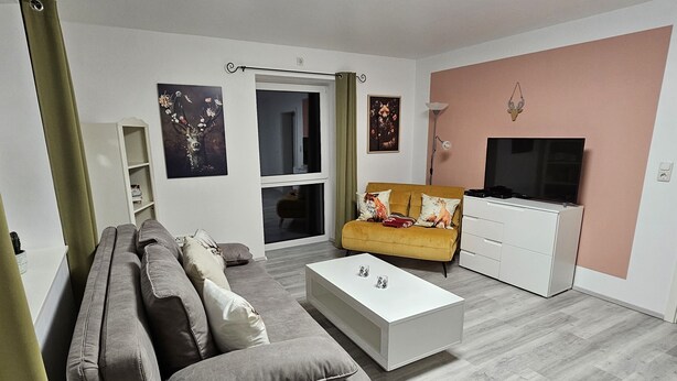 A living room with a grey sofa, a yellow sofa, a white coffee table, and a TV on a stand. - Panoramablick Nahe Oberhof (Geratal)