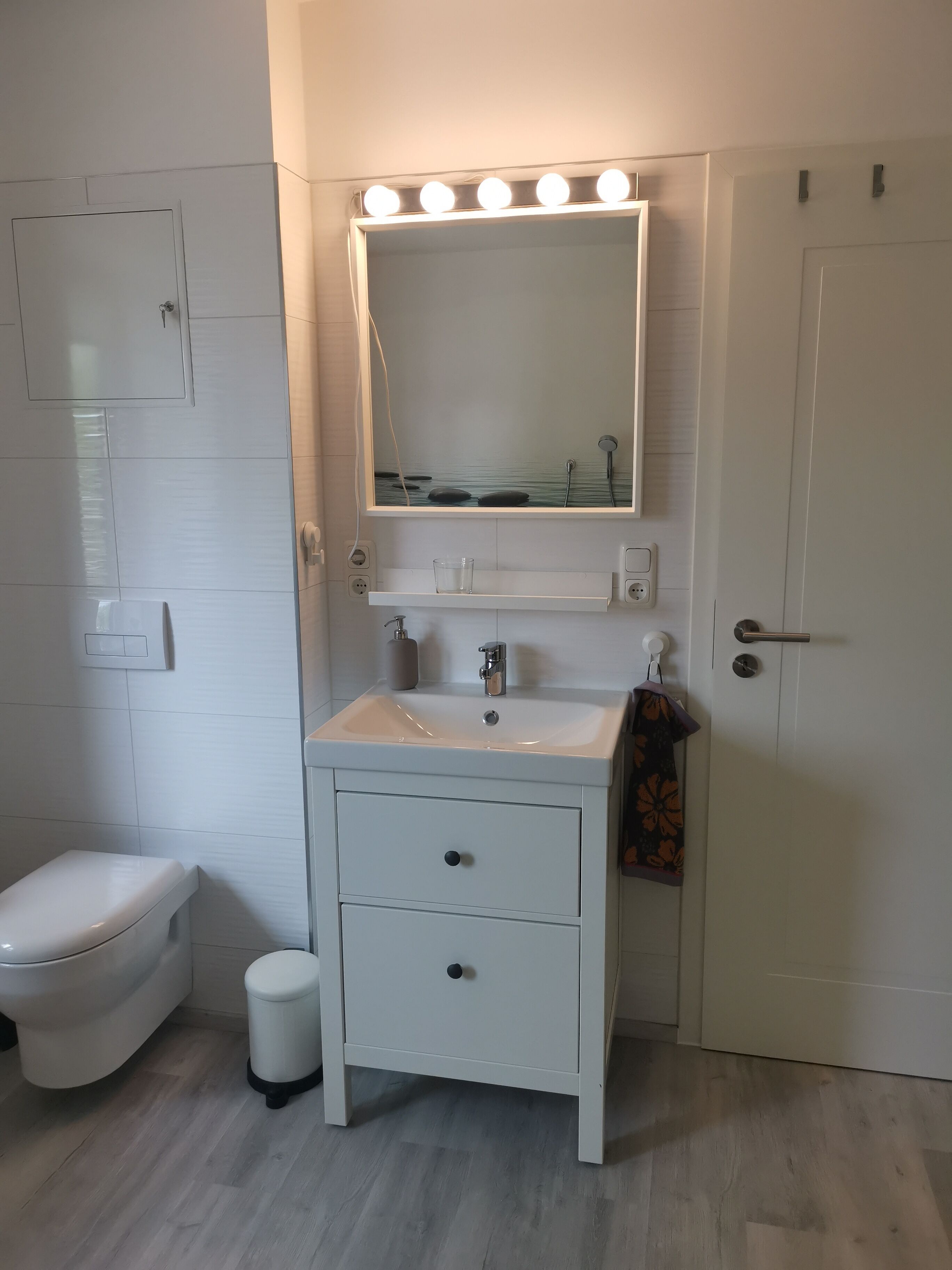 Ein Badezimmer mit weißem Waschtisch, einem beleuchteten Spiegel, einer Toilette und einem Mülleimer.
