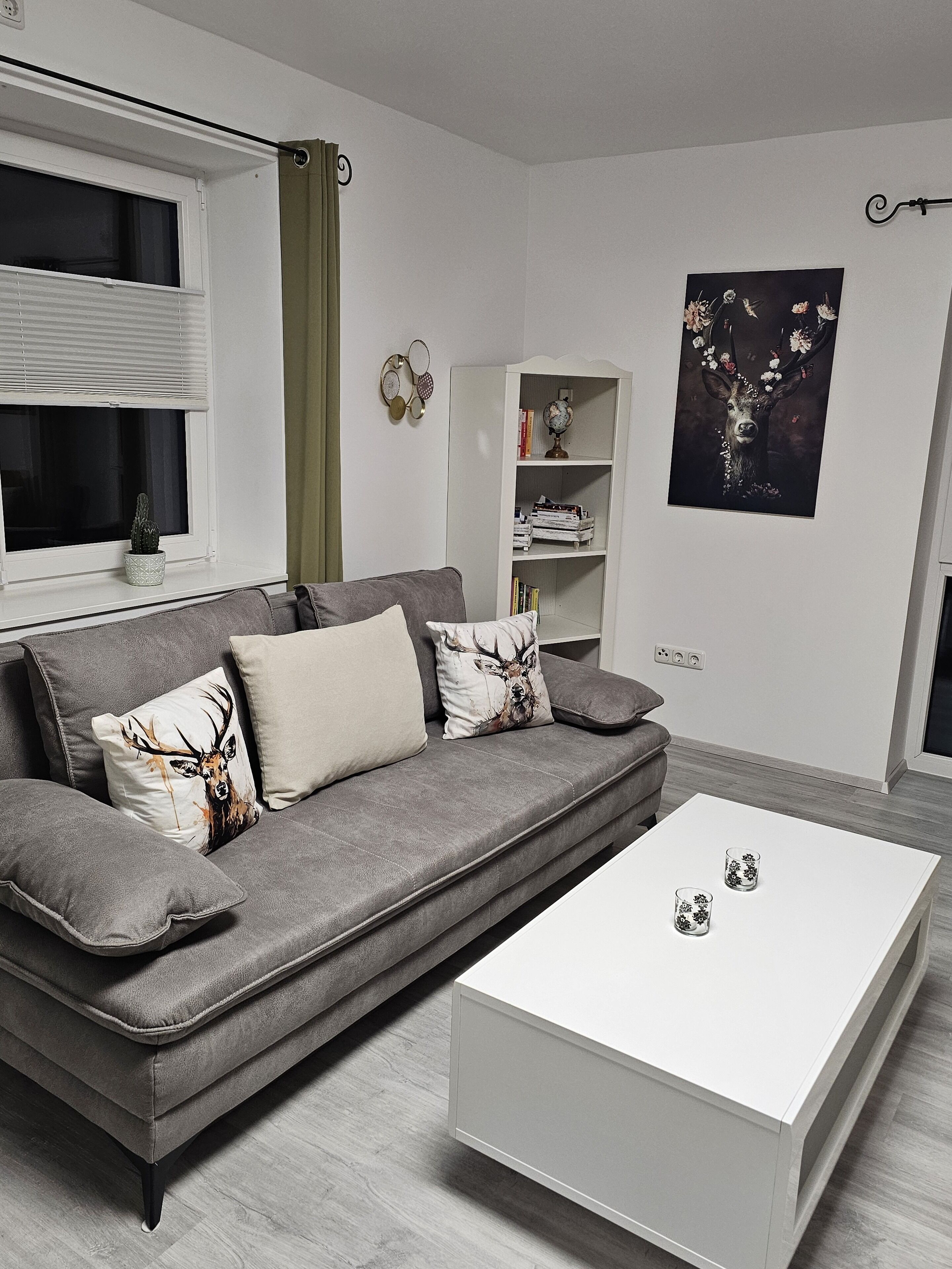 A modern living room with a grey sofa, white cushions, a white coffee table, and a bookshelf with decorative items.