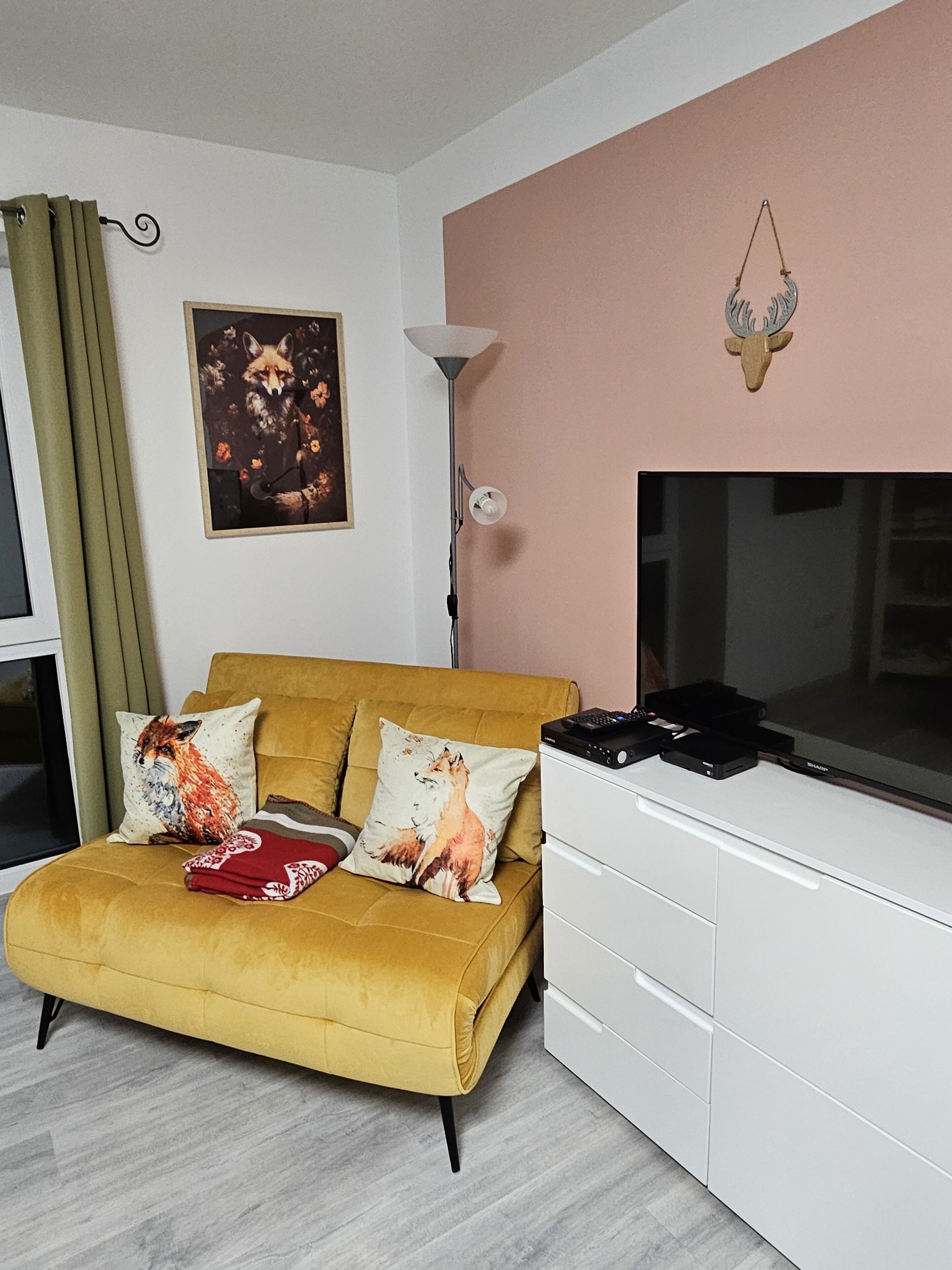 Ein Wohnzimmer mit einem senfgelben Sofa, zwei Kissen mit Füchsen, einem gerahmten Fuchs Gemälde, einer Stehlampe und einem aufgehängten Hirschkopf.