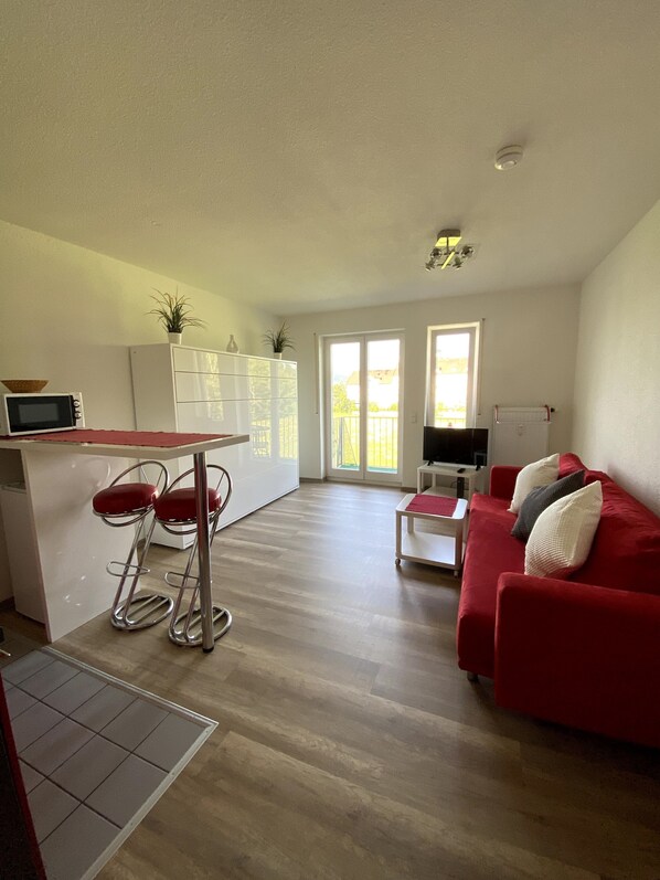 Un espacio habitable moderno con área de cocina, una mesa de comedor con sillas rojas, un sofá rojo con cojines y vistas a través de un ventanal.