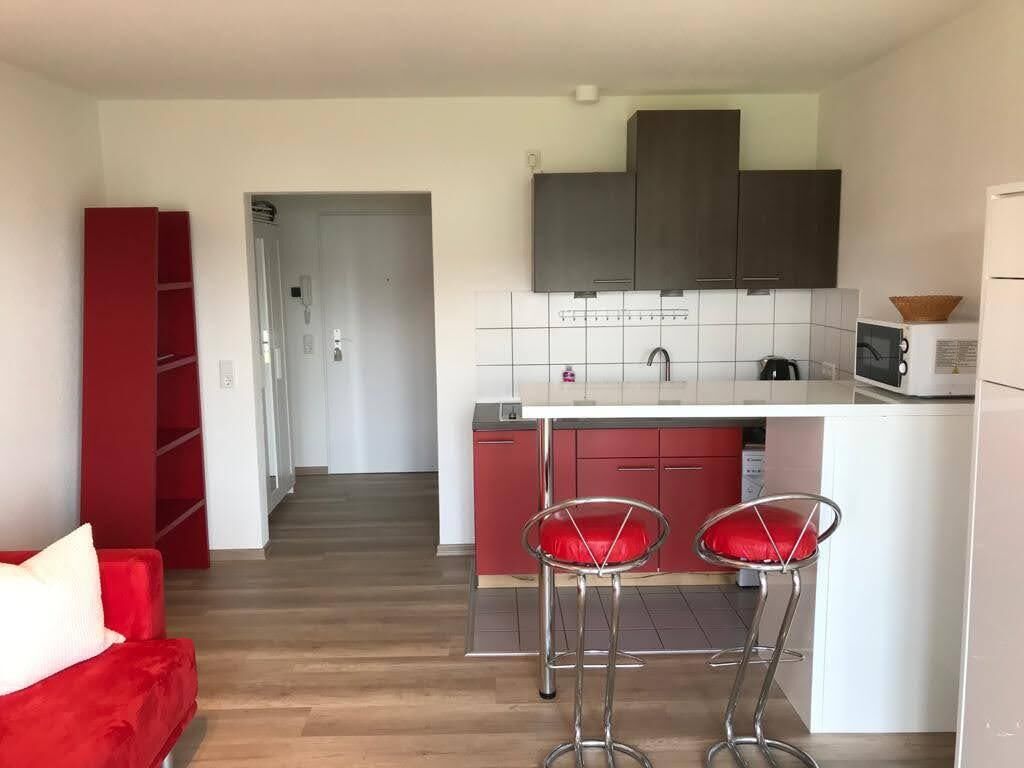 Eine kleine Küche mit weißer Arbeitsplatte, zwei roten Barhocker und dunklen Schränken.