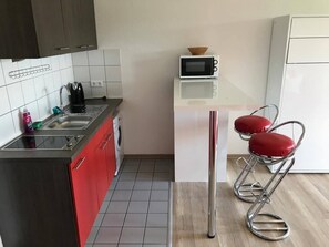 Cocina pequeña con microondas, lavamanos y un taburete rojo.