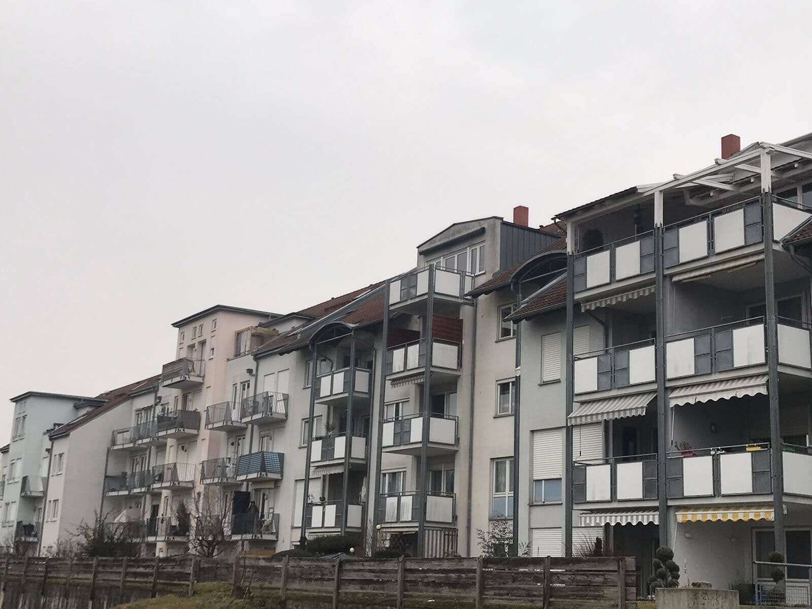 A row of multi-story residential buildings with balconies, a wooden fence in the foreground, and a cloudy sky.