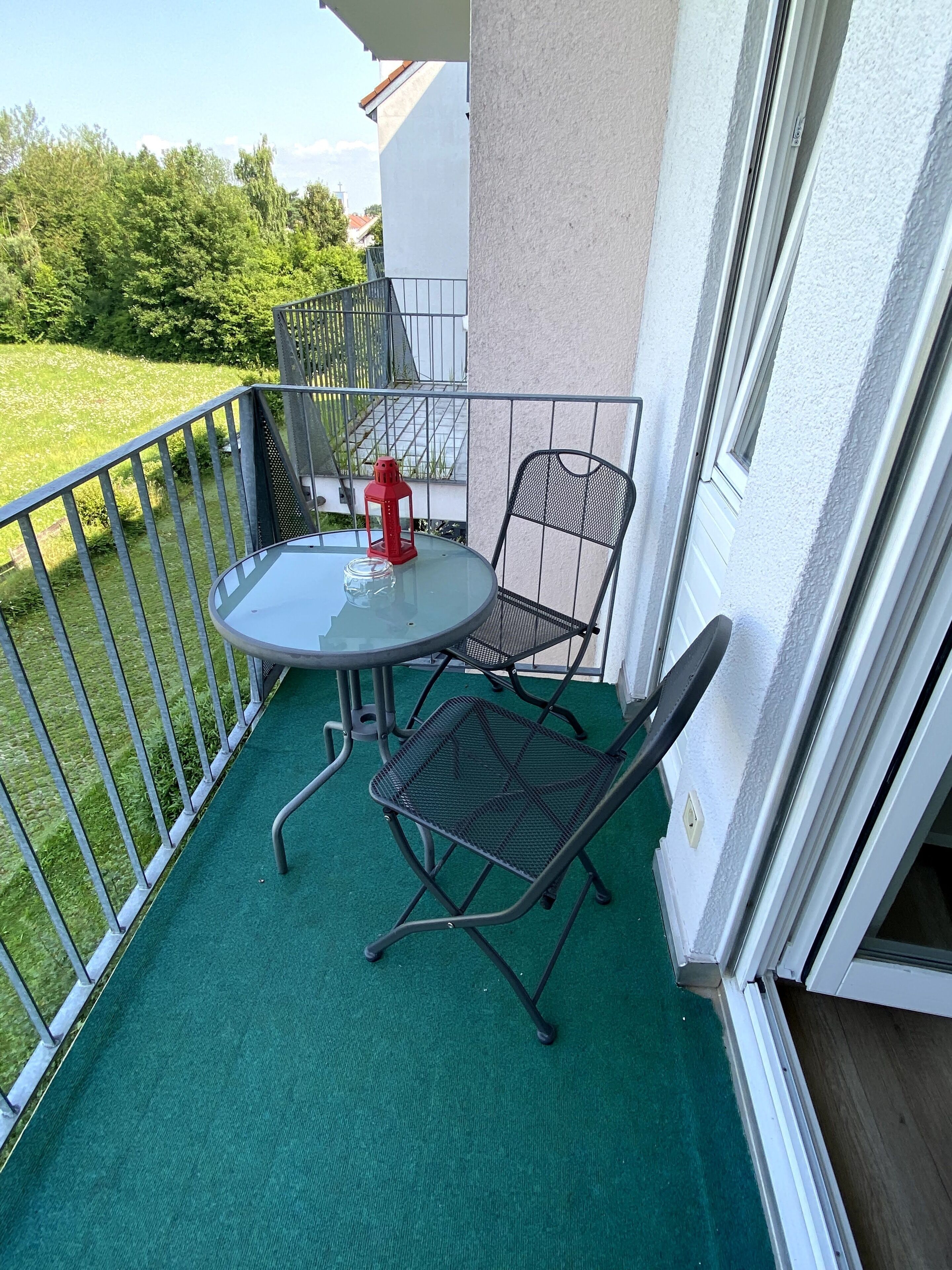 Un balcon avec une table ronde et deux chaises, une lanterne rouge et un sol recouvert d’un gazon artificiel vert.