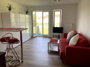 Un salón moderno con un sofá rojo, un televisor sobre un mueble bajo y un balcón con vistas a casas.