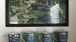 Cuadro enmarco que muestra un paisaje con una edificación tradicional rodeada de vegetación exuberante, sobre una repisa de madera con cuatro tazas de cerámica estampadas.
