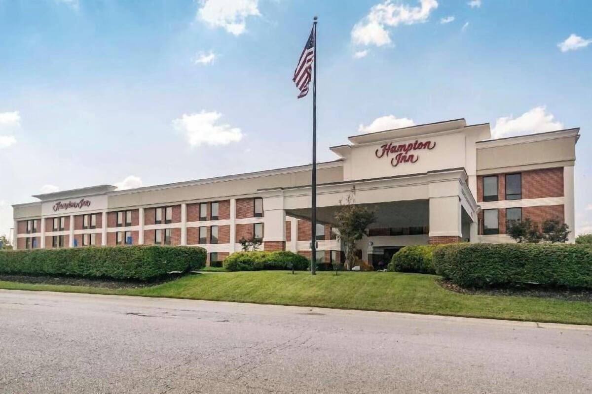 A Hampton Inn hotel with a brick facade, a flagpole with an American flag, and a well-maintained lawn.