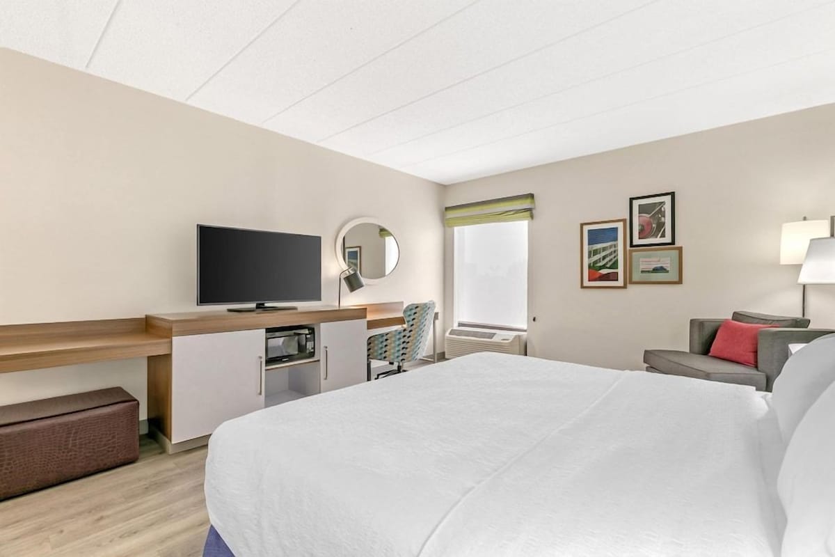 A hotel room with a bed, a desk with a television, a chair, a window, and artwork on the wall.