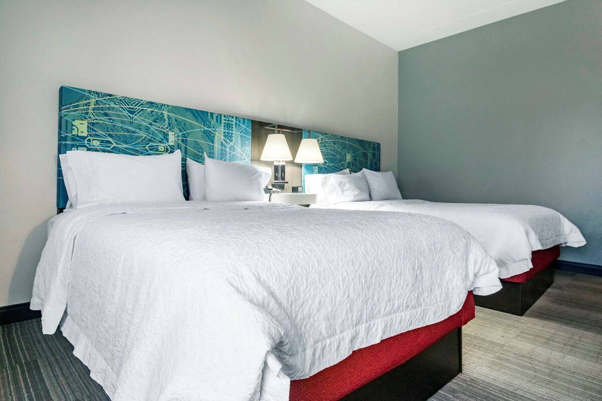 A hotel room with two beds, a headboard with a blue abstract design, two bedside lamps, and a red accent on the lower bed.