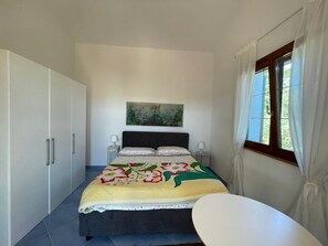 Une chambre à coucher avec un lit, une table de chevet, une chaise, une fenêtre avec des rideaux et un tableau au mur.