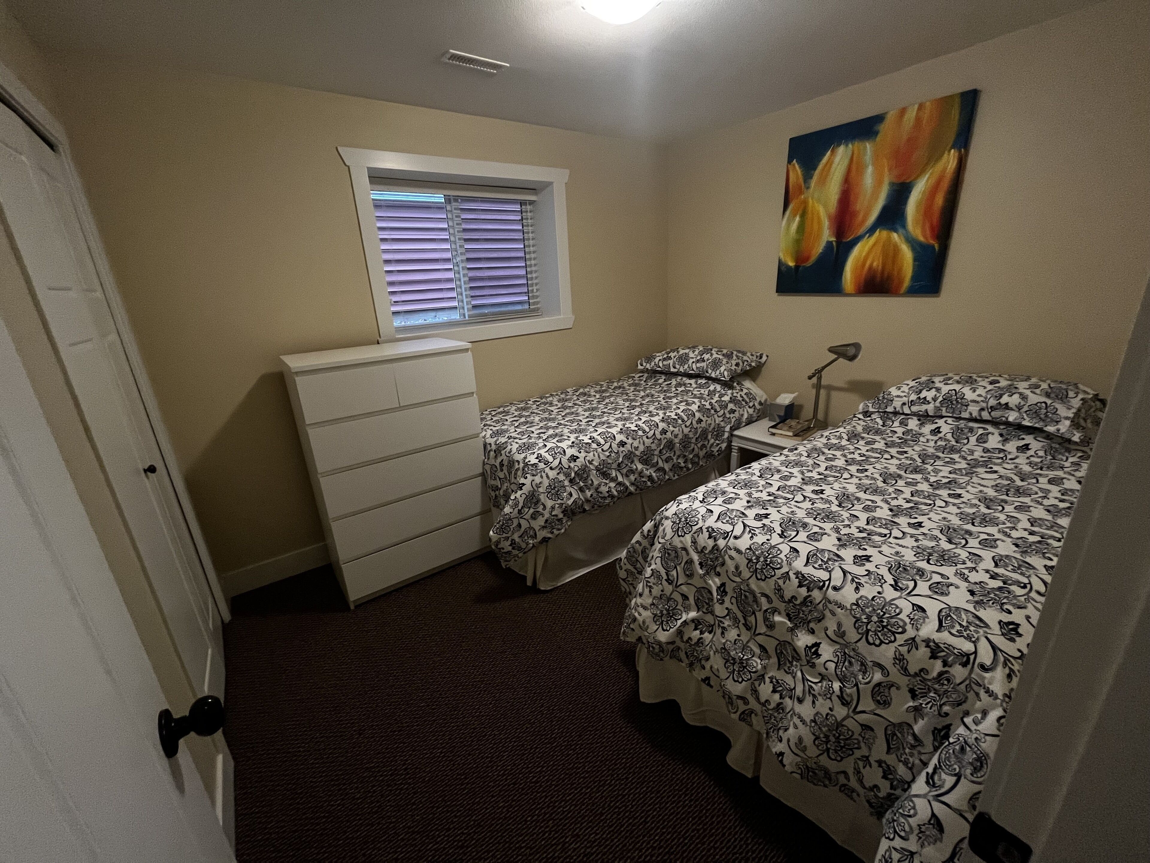 A bedroom with two beds, a dresser, a window with blinds, a painting on the wall, and a carpeted floor.