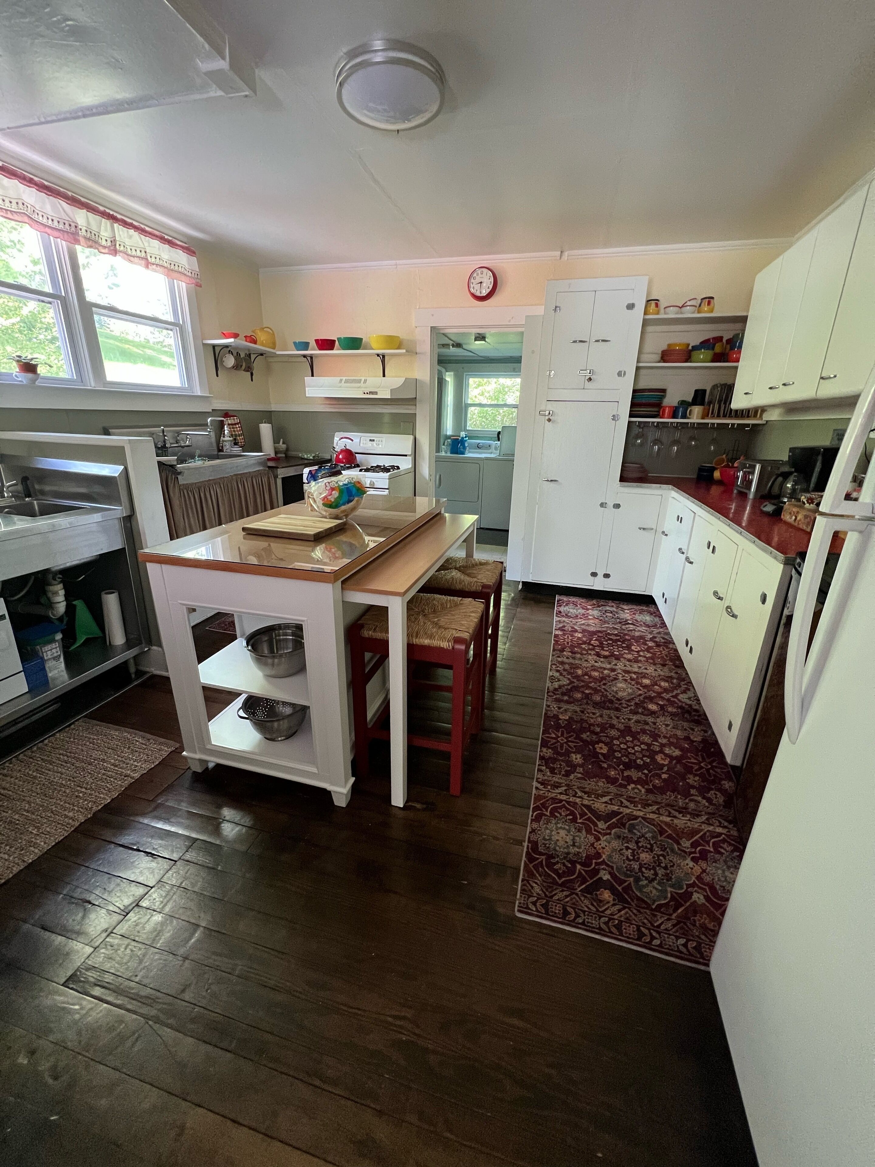 A kitchen with wooden flooring, a dining table with chairs, white cabinets, and a red rug.