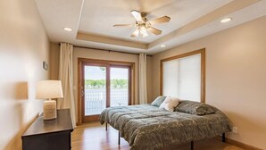 Un dormitorio con cama, ventilador de techo, mesita de noche con lámpara, ventana con cortinas y vista a zonas verdes.