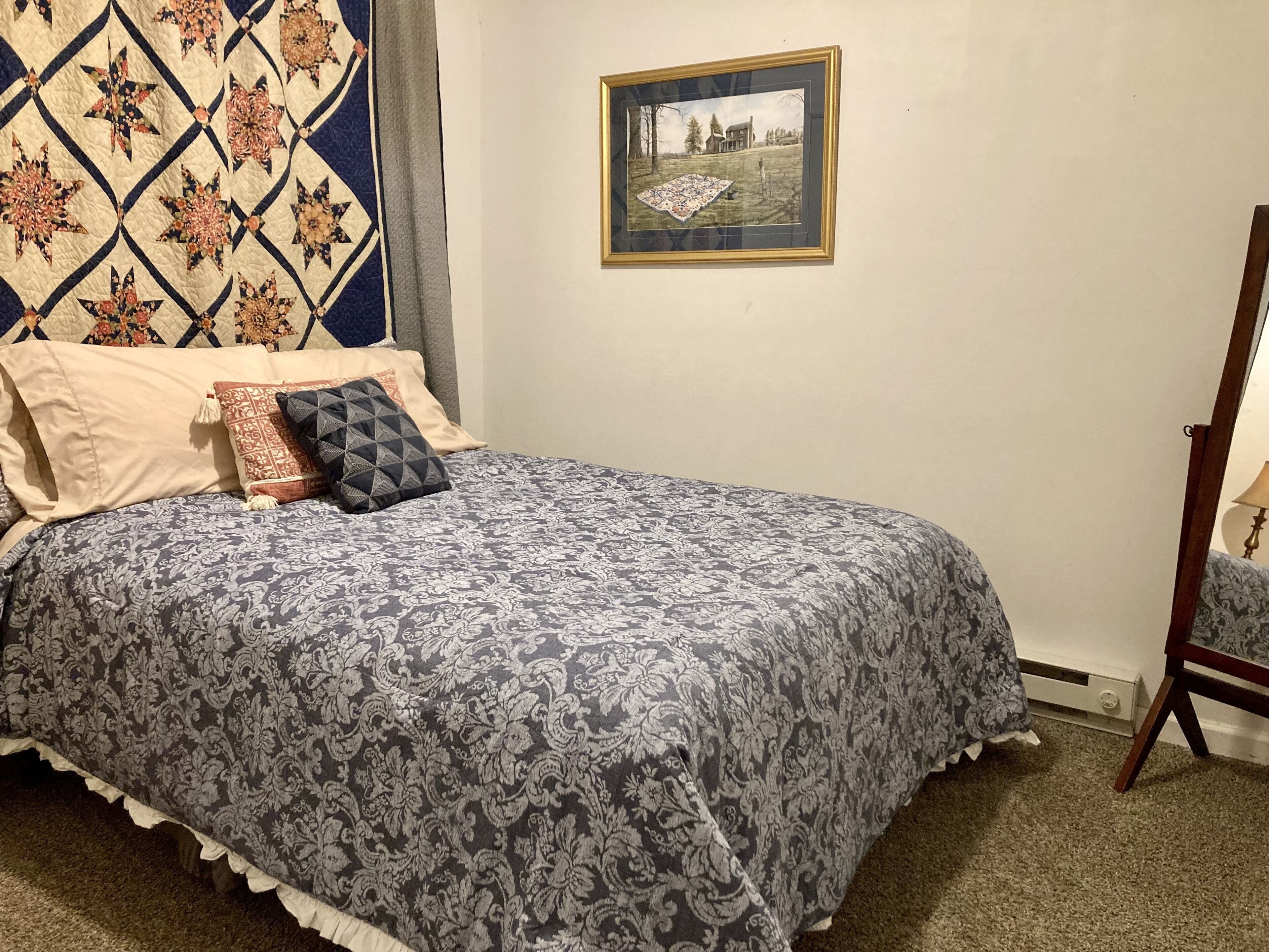 A neatly made bed with a patterned bedspread, a quilt hanging on the wall, and a framed picture on the wall.