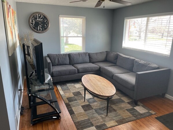 A living room with a gray sectional sofa, a wooden coffee table, a black TV stand, and a large wall clock.