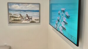 Two framed artworks on a wall: one depicts beach chairs on a sandy shore with waves, and the other shows overwater bungalows connected by a pathway.