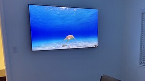 A hotel room with a bed, a flat-screen TV mounted on the wall displaying an underwater scene, and a window with blinds.