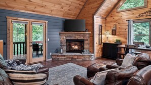 A cozy cabin interior with a stone fireplace, leather furniture, and large windows offering a view of the outdoors. - Stunning Mountain Modern Retreat! 3-Story/Location/Gameroom/Fireplace/Creek (Blue Ridge)