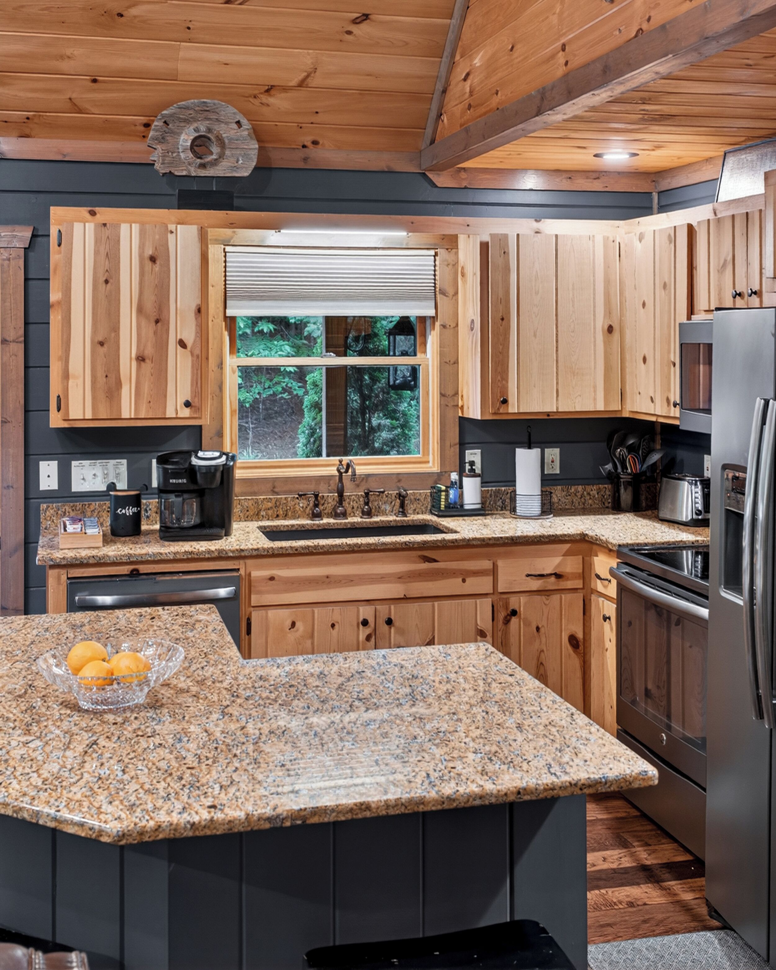 A kitchen with wooden cabinets, granite countertops, a stainless steel refrigerator, and a window with a view of greenery.