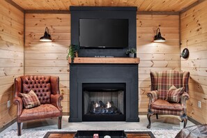 Interior de una cabaña de troncos con chimenea, dos sillones de cuero, un televisor de pantalla plana y paredes de madera.
