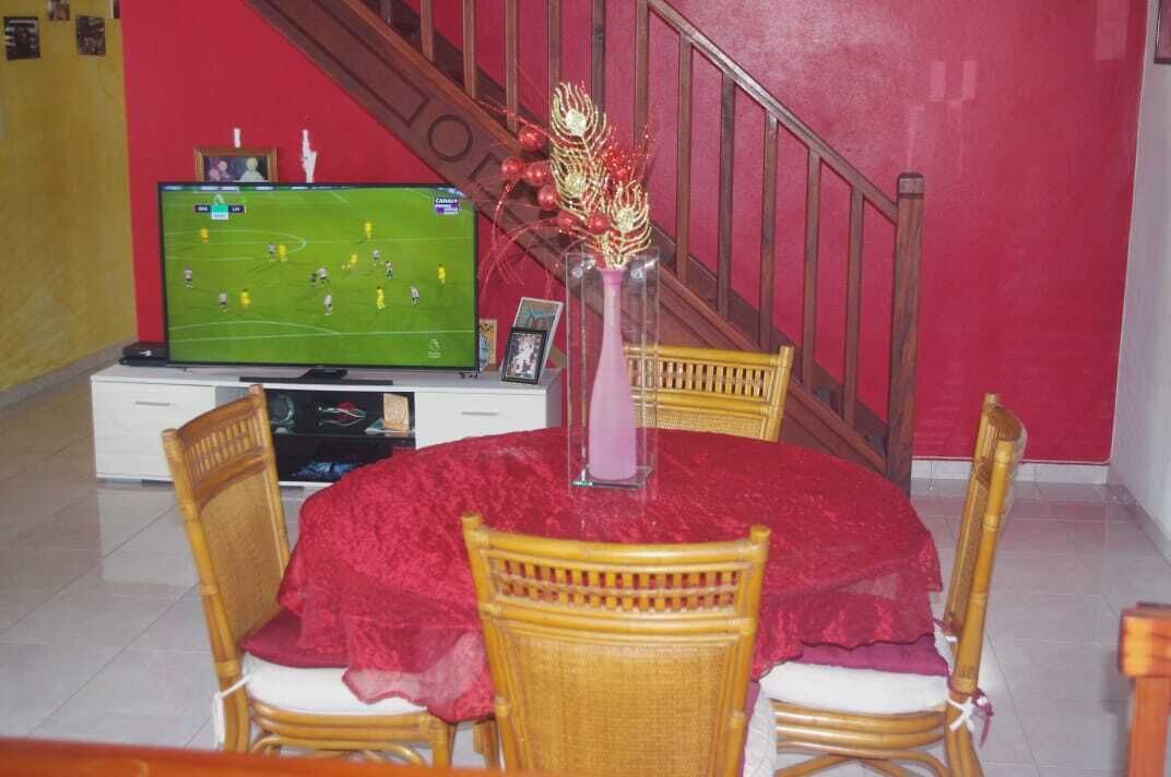 A dining area with a round table covered in a red tablecloth, surrounded by wicker chairs. A television is on a stand to the left, displaying a soccer game.